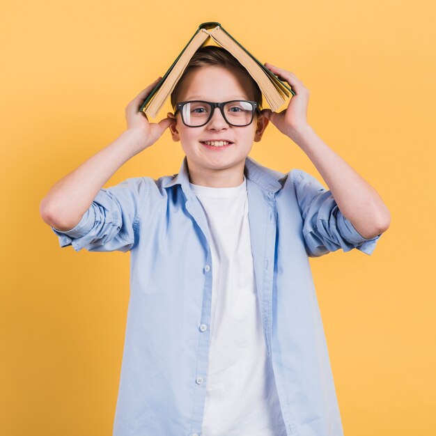 Smiling portrait of a boy holding an open book on his head standing against yellow backdrop