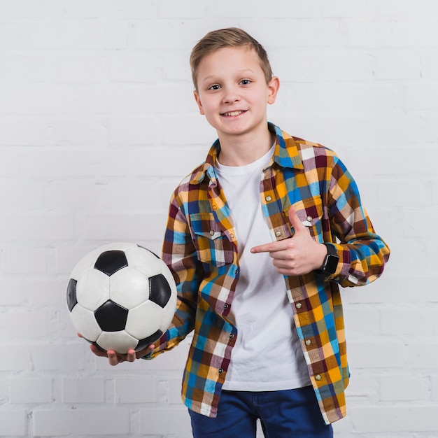 Smiling portrait of a boy showing his soccer ball standing against white brick wall