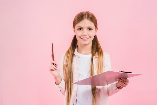 Free photo smiling portrait of a girl holding pencil and clipboard in hands against pink backdrop