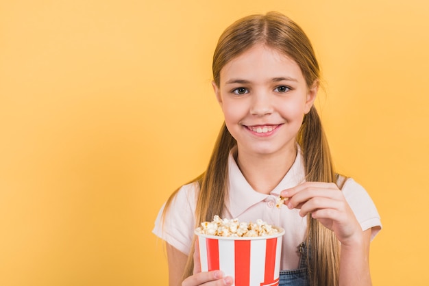 Smiling portrait of a girl holding popcorn bucket against yellow backdrop