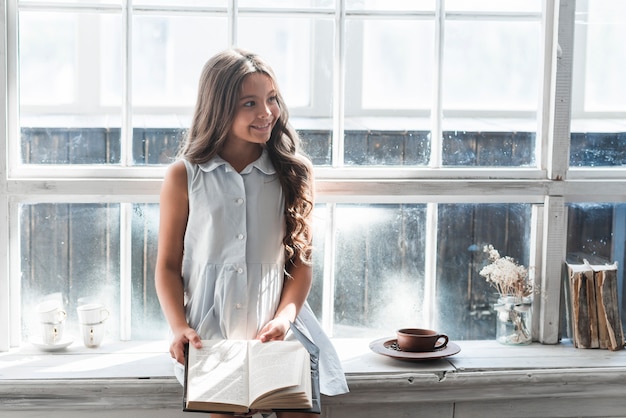 Free photo smiling portrait of a girl sitting on window sill holding book looking away