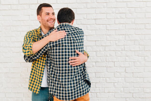 Smiling portrait of a man giving hug to his friend against white brick wall