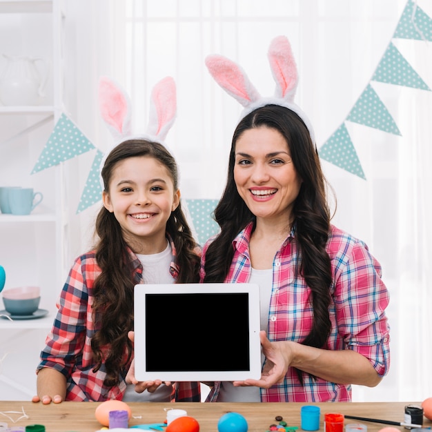 Free photo smiling portrait of mother and daughter showing digital tablet behind the wooden table with easter eggs