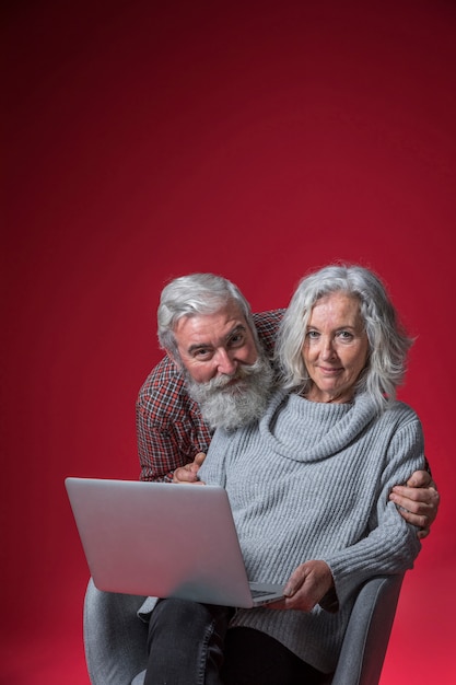 Free photo smiling portrait of a senior man embracing her wife from behind sitting on chair with laptop