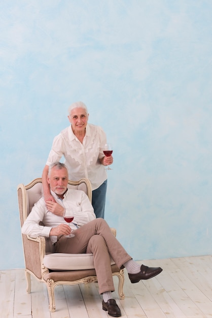 Smiling portrait of a senior man sitting on chair with his wife standing behind holding wine glass