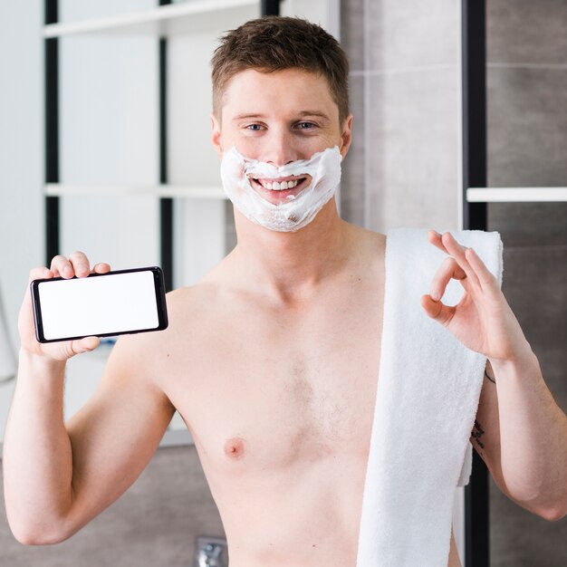 Smiling portrait of a shirtless young man with towel on his shoulder holding smart phone in hand showing ok sign