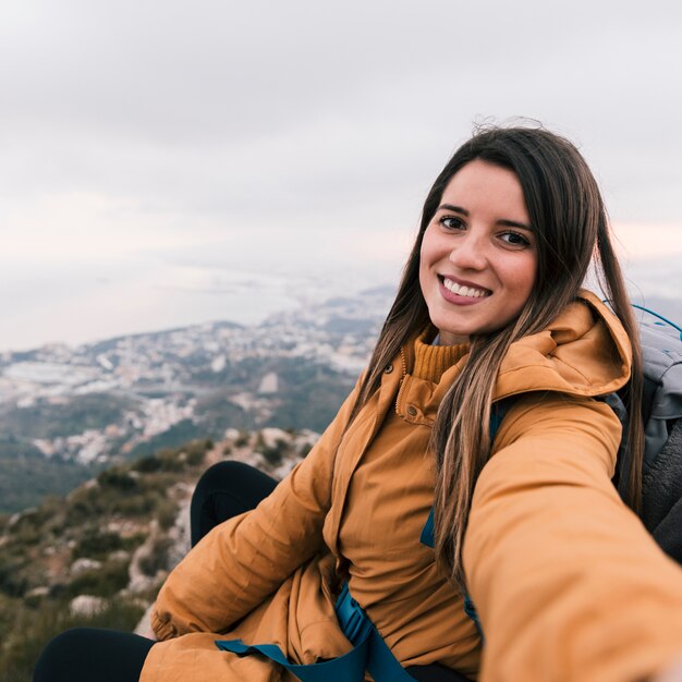 Smiling portrait of a young female hiker taking selfie sitting on mountain top