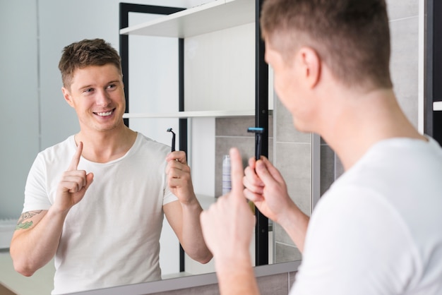 Smiling portrait of a young man holding razor in hand pointing his finger upward looking in the mirror