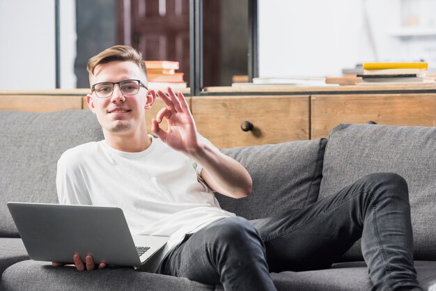 Smiling portrait of a young man lying on sofa holding laptop in hand showing ok sign