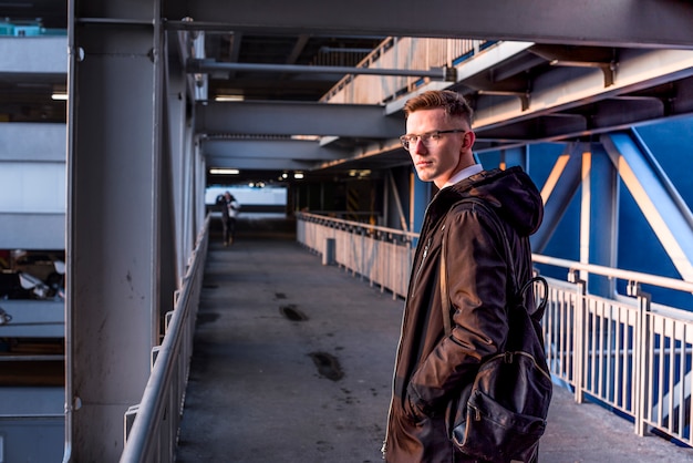 Free Photo smiling portrait of young man with backpack on his shoulder standing on the bridge