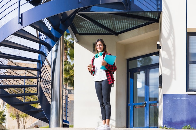 Smiling portrait of a young woman holding books and takeaway coffee cup standing in front of university building