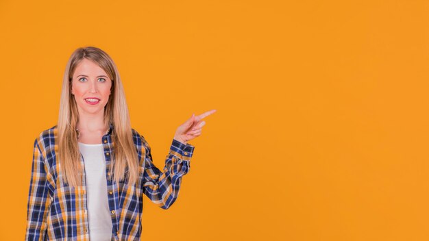 Smiling portrait of a young woman pointing her finger at something on orange background