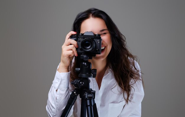 Smiling, positive brunette female in eyeglasses taking pictures with a professional photo camera. Isolated on grey background.