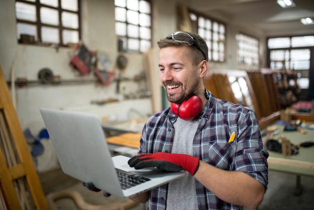 Free photo smiling positive carpenter using laptop computer in carpentry workshop sharing ideas