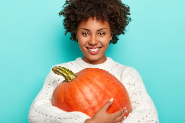 Smiling positive curly haired girl in white sweater, holds autumn pumkin, being in high spirit