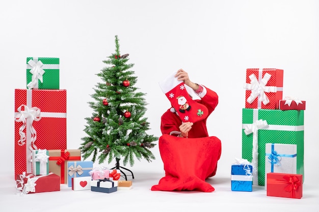 Smiling positive santa claus sitting on the ground and raising christmas sock to his face near gifts and decorated new year tree on white background