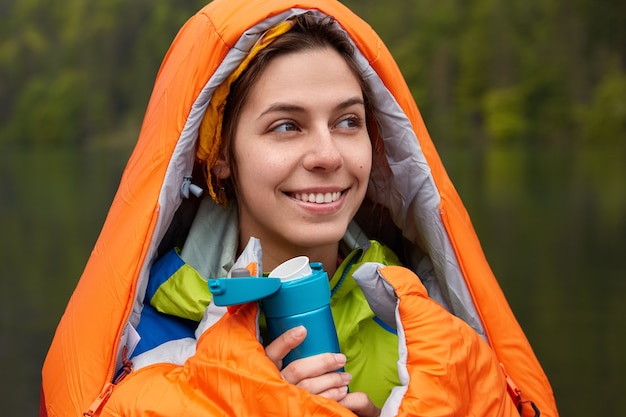 Free Photo smiling positive young female traveler wrapped in sleeping bag, warms with hot drink during cold autumn day