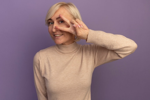 Smiling pretty blonde slavic woman gestures victory hand sign looking at camera through fingers on purple