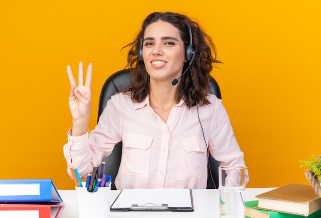 Smiling pretty caucasian female call center operator on headphones sitting at desk with office tools gesturing three with fingers