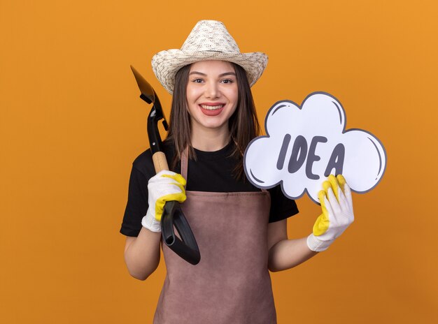 Smiling pretty caucasian female gardener wearing gardening hat and gloves holding idea bubble and spade on shoulder isolated on orange wall with copy space