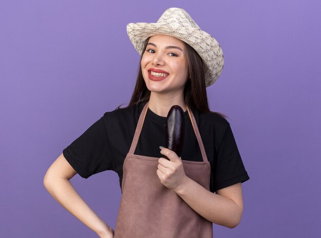 Smiling pretty caucasian female gardener wearing gardening hat holding eggplant on purple