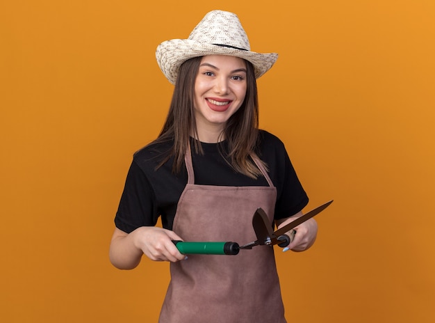 Smiling pretty caucasian female gardener wearing gardening hat holds gardening scissors on orange