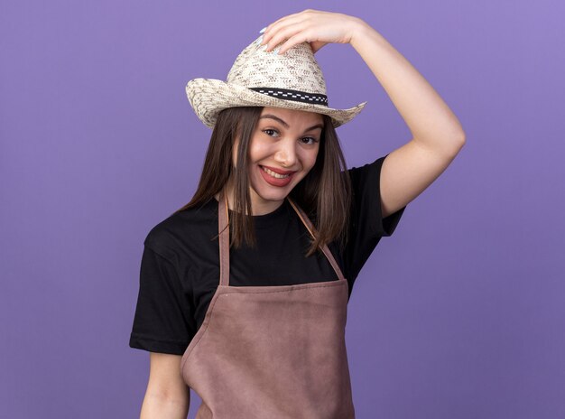 Smiling pretty caucasian female gardener wearing and putting hand on gardening hat on purple