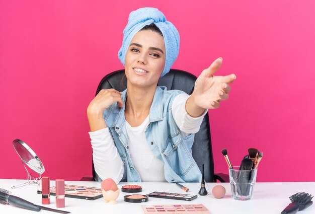 Smiling pretty caucasian woman with wrapped hair in towel sitting at table with makeup tools stretching out her hand isolated on pink wall with copy space