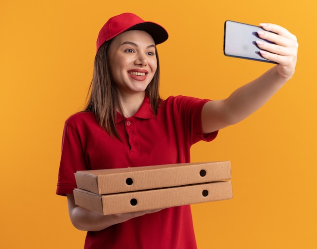 Free photo smiling pretty delivery woman in uniform holds pizza boxes and looks at phone taking selfie