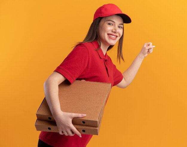 Smiling pretty delivery woman in uniform keeping fist and holding pizza boxes on orange