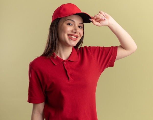 Smiling pretty delivery woman in uniform puts hand on cap isolated on olive green wall with copy space