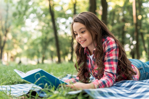 Smiling pretty girl reading book while lying on checkered blanket at park