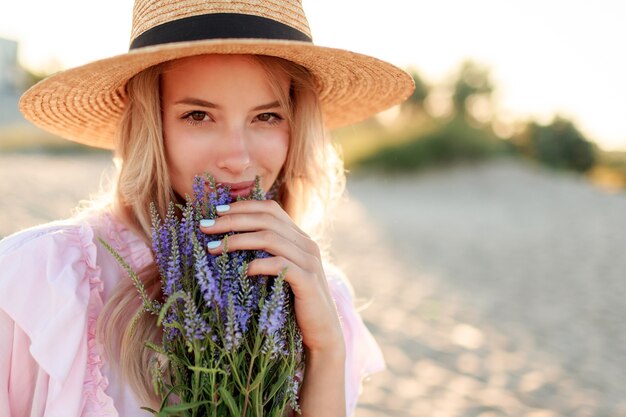 Smiling pretty girl in  straw hat posing on sunny beach near ocean with bouquet of flowers. Close up portrait.