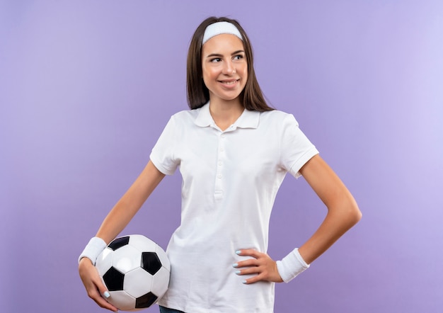 Free Photo smiling pretty sporty girl wearing headband and wristband holding soccer ball looking at side with hand on waist isolated on purple space