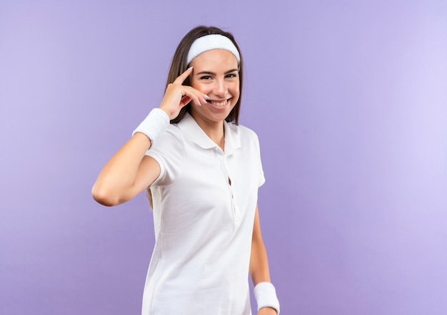 Smiling pretty sporty girl wearing headband and wristband putting finger on temple isolated on purple space 