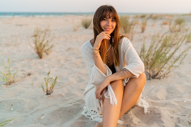 Free Photo smiling pretty woman in bohemian summer outfit with stylish necklace sitting on sand