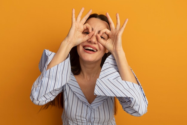 Smiling pretty woman looks at front through fingers isolated on orange wall