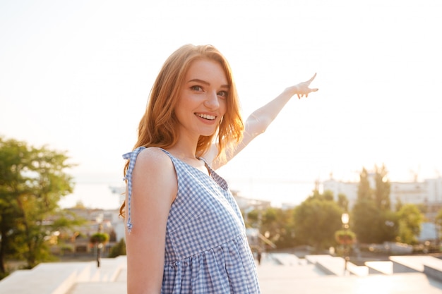 Smiling pretty woman pointing finger away while standing outdoors