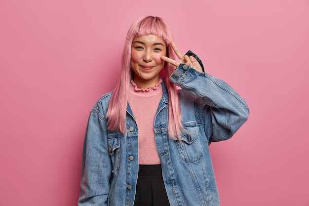 Smiling pretty woman wishes luck and peace, shows victory sign, makes cute seducing face, has fun, long dyed straight pink hair, wears jean jacket