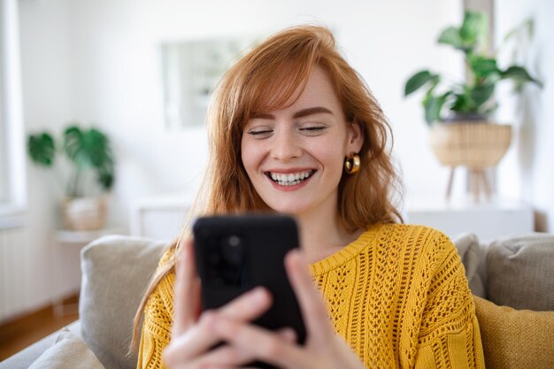 Smiling pretty young woman relaxes on the sofa in her living room while using her mobile smart phone for social media and surfing the internet