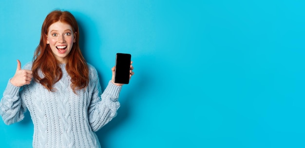 Free photo smiling redhead girl showing smartphone screen holding phone and demonstrating app making thumb up i