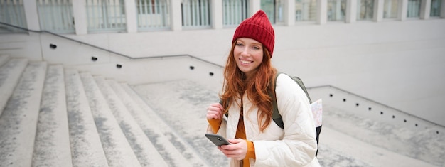 Free Photo smiling redhead girl student tourist walks around city goes up the stairs looks at mobile phone map