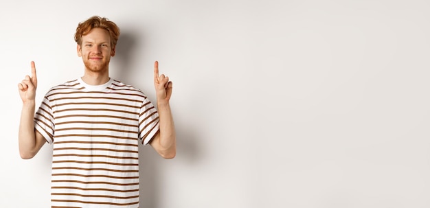 Free Photo smiling redhead guy with beard pointing fingers up and showing advertisement standing over white bac