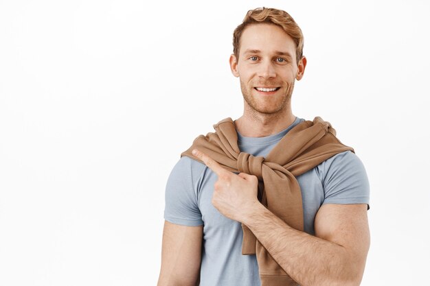 Free photo smiling redhead man with muscles big biceps, pointing at upper left corner, showing advertisement on product on copy space, standing over white wall