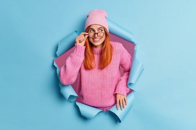 Smiling redhead schoolgirl focused happily aside with thoughtful face expression keeps hand on spectacles wears hat and pink sweater.