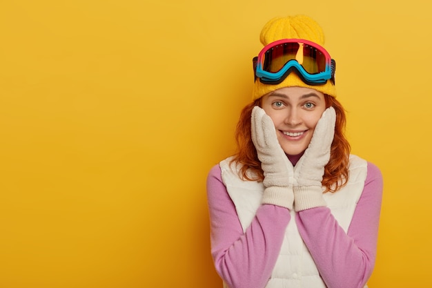 Free photo smiling redhead snowboarder girl has happy expression, touches cheeks, isolated over yellow background