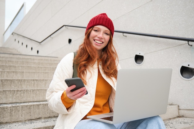 Smiling redhead woman with mobile phone and laptop sitting on stairs outside building connects to pu