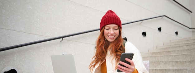 Free photo smiling redhead woman with mobile phone and laptop sitting on stairs outside building connects to