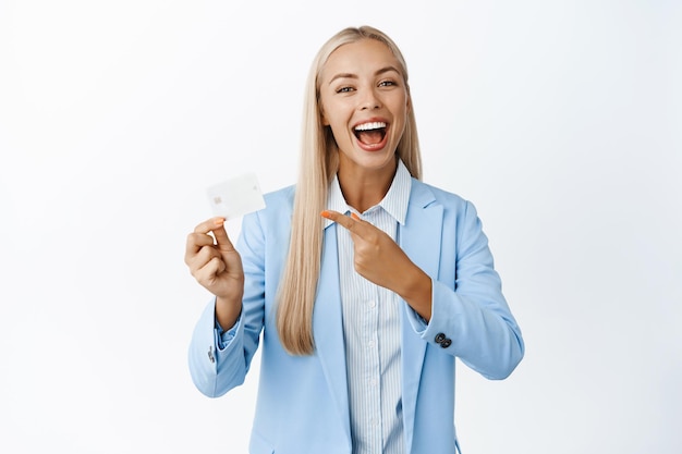 Smiling saleswoman pointing at credit card advertising bank standing in suit over white background Finance concept