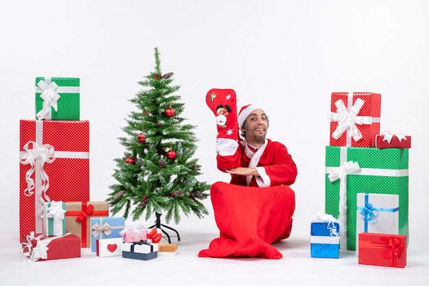 Smiling santa claus sitting on the ground and wear christmas sock near gifts and decorated new year tree on white background